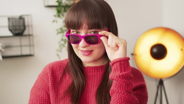 Sassy caucasian young adult woman looking at camera and touching her pink sunglasses while standing in modern apartment. High quality 4k footage