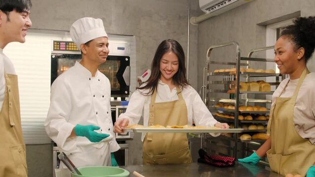 Young Asian female cooking class student shows tray of baked pies from oven, good smell for group of chefs, happy pastry cuisine in culinary course lesson, food occupation in stainless steel kitchen.