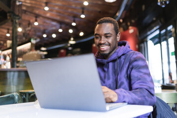 Fototapeta premium Positive African American male freelancer sitting at table and typing on netbook while working remotely in modern cafe
