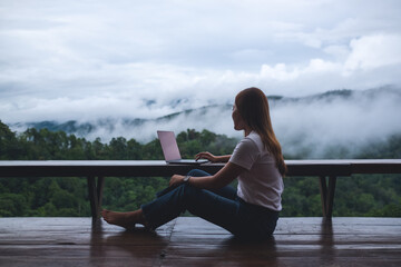 Fototapeta premium A young woman using and working on laptop computer while sitting on balcony with a beautiful nature view on foggy day