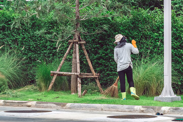 Female worker cleaning up leaves with traditional broom tool in ornamental garden. Woman wear rubber gloves with hat and boots sweeping away the fallen leaves. Wooden Broom in hand sweeping up ground.