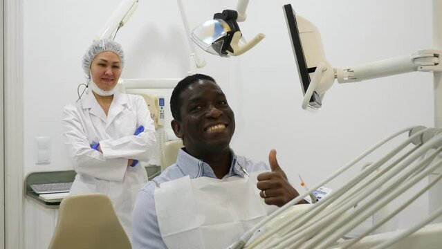 African-american man patient sitting on dental chair and smiling. Asian woman dentist standing in background.