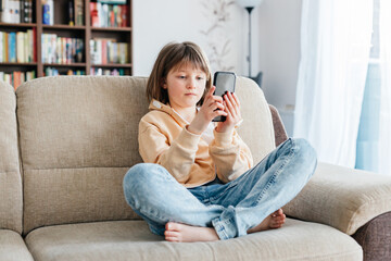 Fototapeta premium Schoolgirl is sitting on the sofa and playing mobile phone or chatting with friends. Children use technology. Modern gadgets and teenagers.