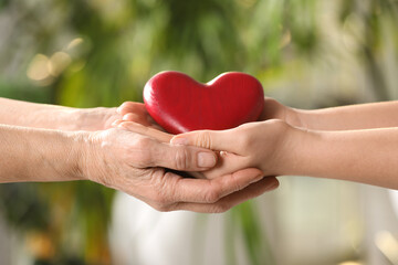 Fototapeta na wymiar Young and elderly women holding red heart in hands on blurred green background, closeup