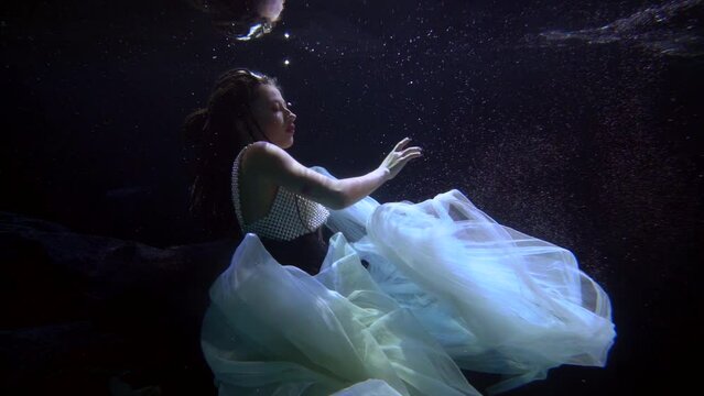 a woman in a lush light dress hovered in the dark water with bubbles and beautifully moves hands. profile view