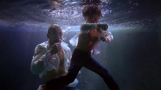 a man and a boy in unbuttoned white shirts are boxing in dark water with bubbles