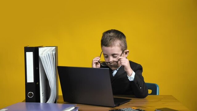 A small confident businessman boss sits on a yellow background with papers and a laptop. The boy was very tired of working. Children as adults