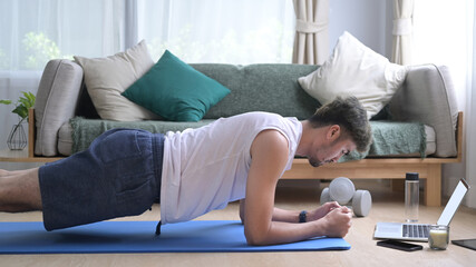 Young man in sports clothes doing plank on blue mat and watching fitness lessons online on laptop. Sport at home, workout, fitness and exercises