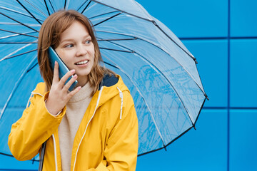 young caucasian woman dressed in a yellow raincoat holds a transparent umbrella and uses a smartphone