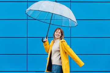 portrait of a pretty girl dressed in yellow raincoat posing while standing with an open umbrella over blue background