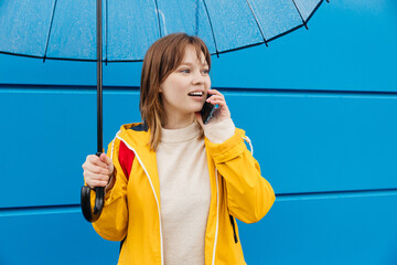 young caucasian woman dressed in a yellow raincoat holds a transparent umbrella and talking on a mobile phone