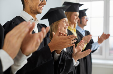 Happy graduates in hats and robes standing in row and applauding. Cheerful male and female...