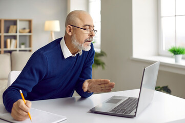 Focused bald senior man in glasses studying at home, sitting at table with modern laptop computer,...