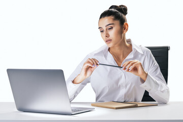 Business process and job concept with pretty young woman sitting at white desk, looking at laptop screen and carrying pencil in hands on abstract white wall background