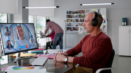 Side view of mature businessman in headphones having video conference with colleagues on computer
