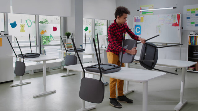 African-American teen boy clean class after lesson putting chairs on desks and sweeping floor