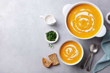 Pumpkin, vegetable cream soup in white pot and bowl. Grey background. Copy space. Top view.