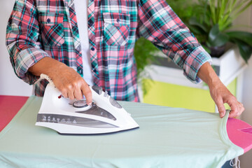 Closeup of mature woman in checkered shirt ironing clothes at home on ironing board