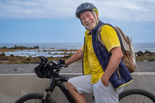 Smiling bearded cyclist senior man along the sea beach carrying a backpack, wearing helmet running with electro bicycle looking at camera. Concept of healthy and sustainable mobility