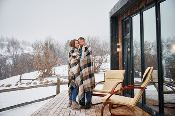 Full length of man and woman wrapped in blanket standing near chairs outside wooden house with panoramic windows. Happy couple hugging outdoors under winter snow.