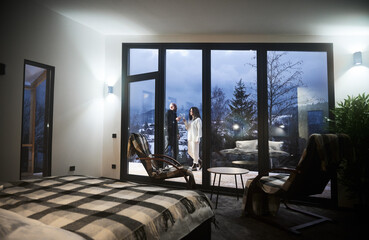 Beautiful view of man and woman toasting with wine behind panoramic window of scandinavian house barnhouse. Interior of wooden house room with chairs, bed and couple in love outside building.