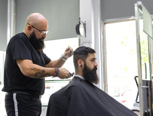 side view shot of bald male barber shaving a client with an electric razor into his place of work