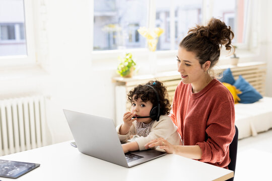 Little girl with headset sitting on lap of mother using laptop