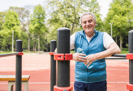 Smiling senior man holding water bottle standing amidst gymnastics bar
