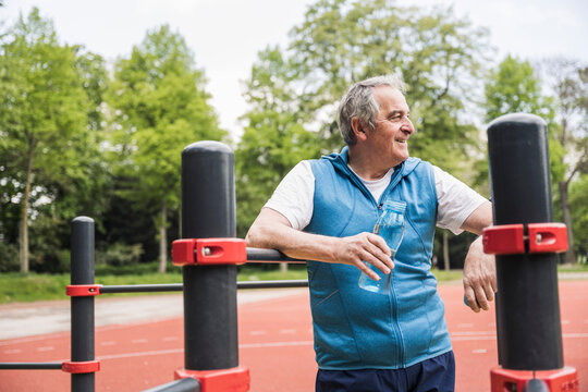 Happy senior man with water bottle standing by gymnastics bar