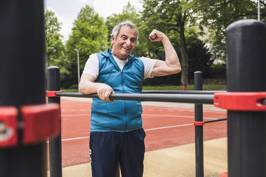 Happy senior man flexing muscles standing by gymnastics bar