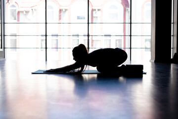 silhouette attractive caucasian yogi woman standing in balasana pose exercising alone in the gym against the background of a window dressed in sportswear, healthy lifestyle concept,
