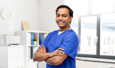 healthcare, profession and medicine concept - happy smiling doctor or male nurse in blue uniform with crossed arms over medical office at hospital background