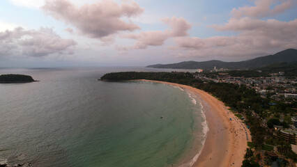 View of the beach and the island from a height. Sunset. People walk on the beach, relax. There are clouds in the sky. Green hills on the island of Phuket. There are hotels, lanterns are lit. Journey