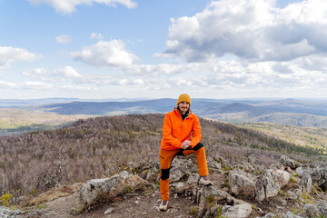 A guy in orange clothes stands on top of a mountain, a happy tourist traveler, a man smiling, a climber at the peak of the mountains, the happiness of a man, a hike