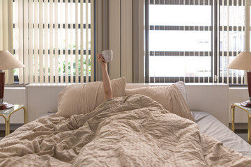 girl hiding under duvet sheets in bed holding cup with arm raised - difficult morning waking up - need of coffee - hotel room holiday and hangover after-party concept - obrazy, fototapety, plakaty