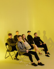 Group of six young boys and girls sitting on armchairs on a yellow background with yellow light and thinking