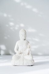 Decorative white Buddha statuette on the white background with sun light shadows. Meditation and relaxation ritual. Buddha birthday. Minimalism. Copy space. Vertical card. Selective focus.