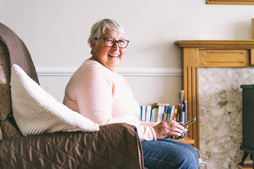 Portrait of happy laughing senior woman using phone while sitting on armchair at home. Mature older...