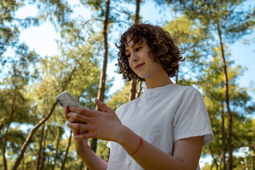 Outdoor portrait of a young redhead woman holding phone on hands or using app or messaging with friends on mobile phone while standing green city park, outdoors.