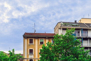 Behind the spring green maple leaves, an old yellow stone residential building with windows and shutters, a green rooftop terrace against a cloudy sky in the early morning in Milan, Italy