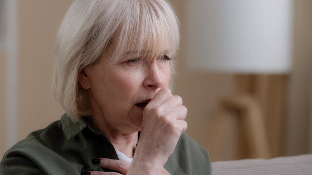 Portrait old woman coughing at home. Close up unwell Caucasian aging mature lady coughs having pain in chest coronavirus symptoms. Grandmother suffer from sore throat cold flu virus cough infection