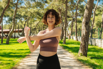 Portrait of a smiling fitness redhead woman stretching arms on city park, outdoors. Fitness woman exercise warm up stretch arms before workout.
