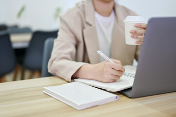 Professional businesswoman using laptop computer, taking notes and sipping coffee.
