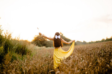 A girl with long hair and a white bow stands in a yellow skirt in a field with spikelets