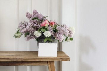 Springtime still life. Blank horizontal greeting card, invitation. Vase with bouquet of lilac, viburnum and tulips on old wooden table. White wooden wall background. Empty copy space. Rustic interior.