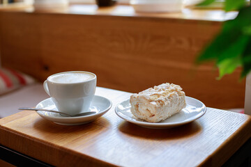 a cup of fresh cappuccino and a meringue roll on the table in a coffee shop. close-up
