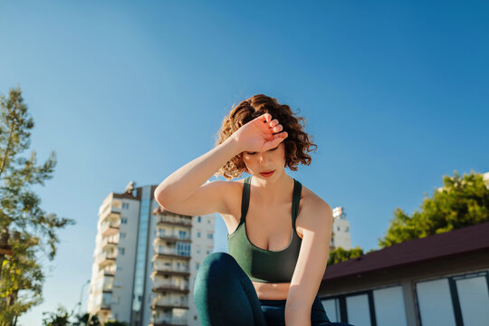 Cute Redhead Woman Sitting On Gras Wearing Sports Bra And Blue Yoga Pants After Jogging Catching Her Breath, Wiping Her Sweat From Her Forehead.