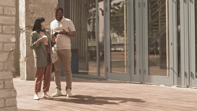 Fill length slowmo of cheerful young diverse business couple in smart casual outfits having conversation while walking at downtown on warm sunny day, using navigation app on smartphone