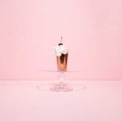Golden paper cup filled with whipped cream and cherry on the top on glass cake stand against pink background. Summer cupcake. Minimal concept.