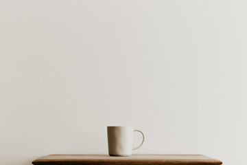 A beige ceramic mug on a brown wooden board in front of a white wall. Hand made coffee cup with...